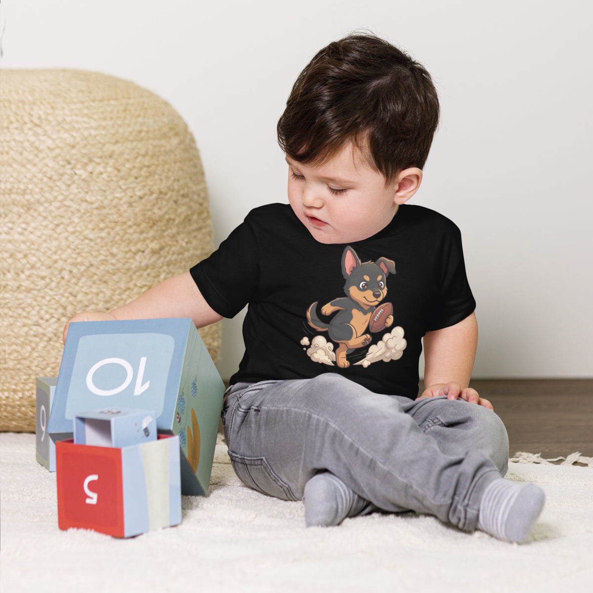 Toddler boy sitting by toy blocks wearing a black 'football pup' toddler t-shirt, puppy running with a football, cozy bluesky
