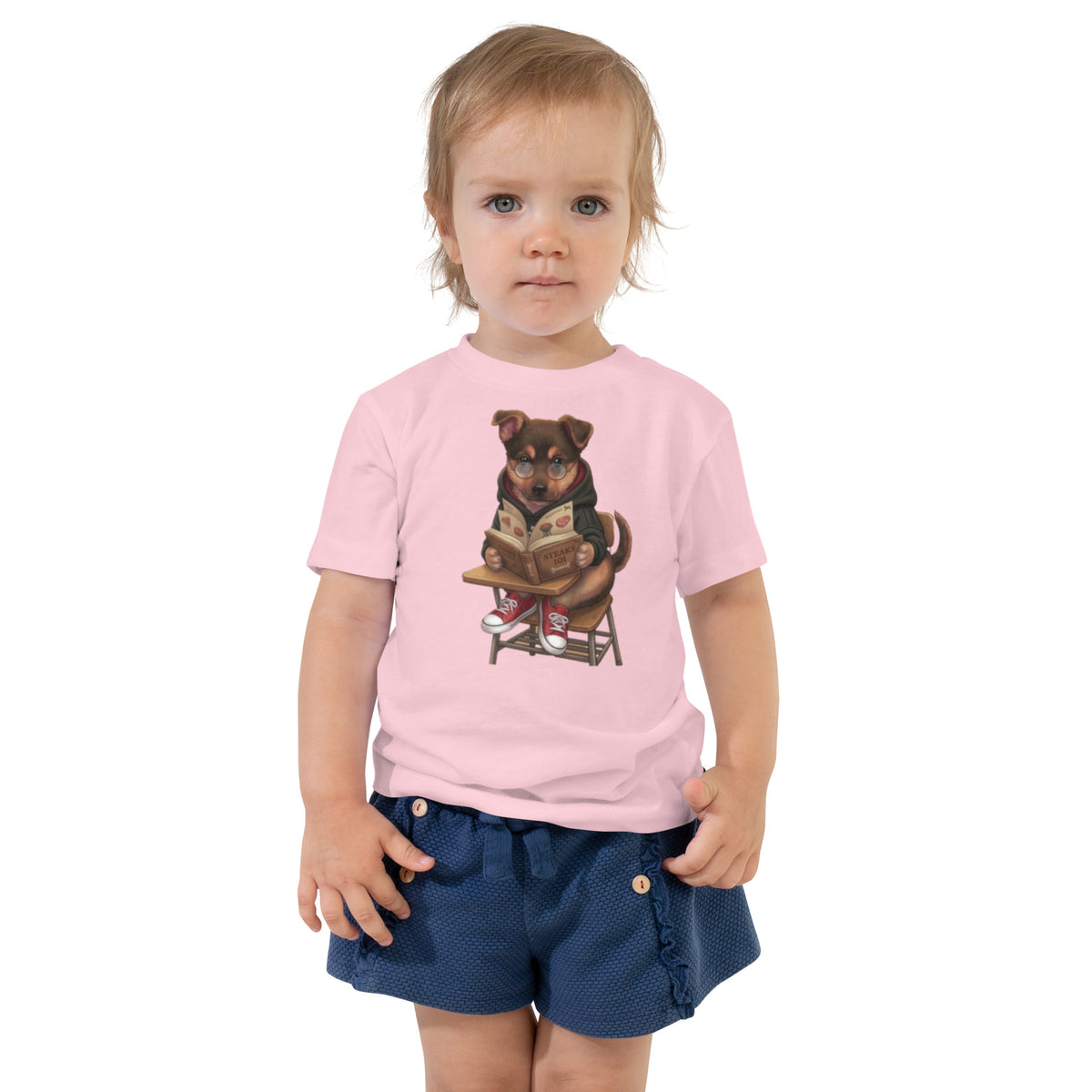 Toddler girl in navy shorts wearing a pink 'the studious pup' toddler t-shirt, dog reading a book at a desk, cozy bluesky