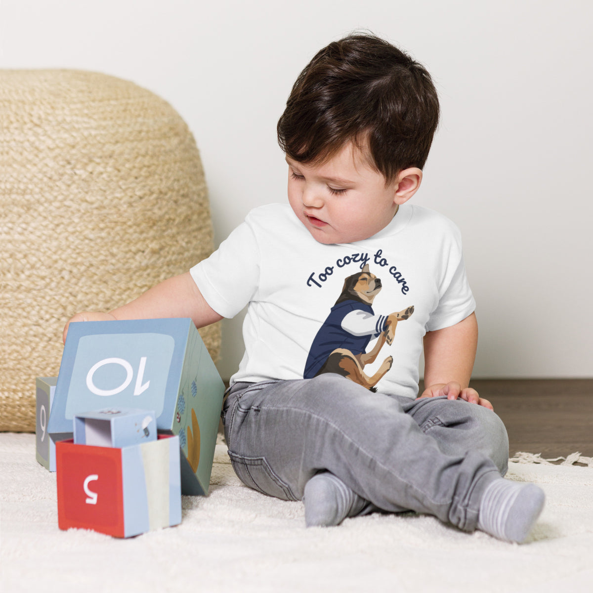 Seated toddler boy wearing a white 'too cozy to care' toddler t-shirt, dog in a navy jacket, too cozy to care, cozy bluesky